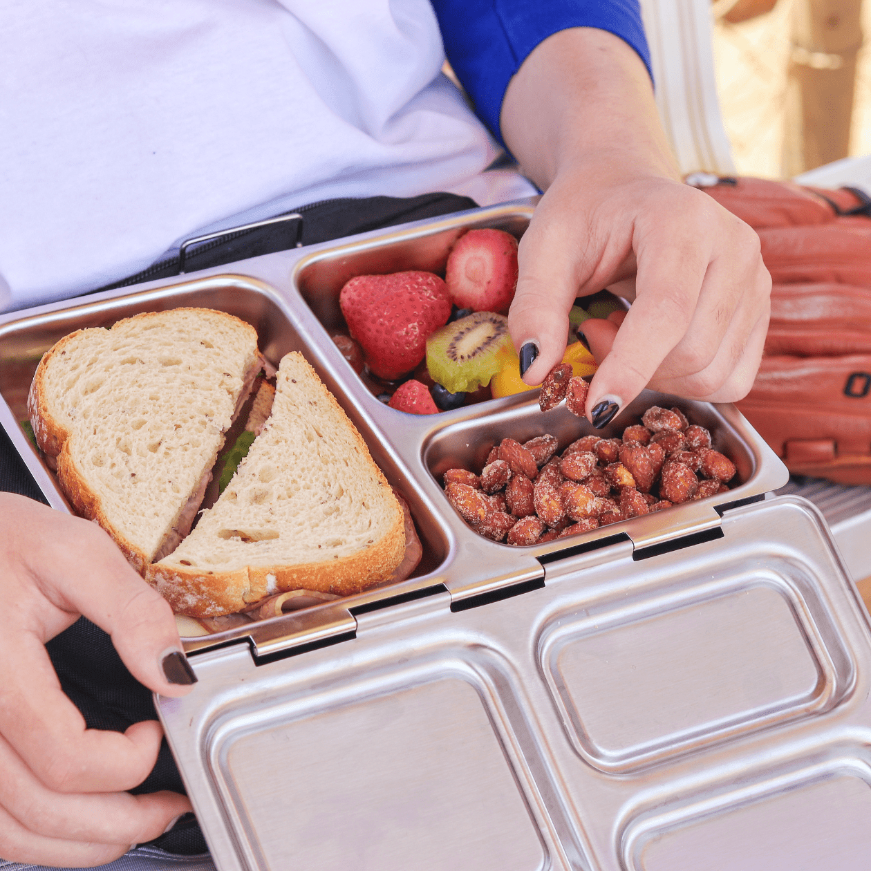 Close up of teenager eating out of Launch lunchbox at a baseball field. Box is filled with nuts, fruit and a sandwich