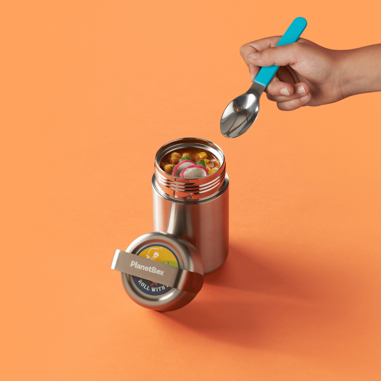 Hand holding spoon going to take a scoop of tortilla soup from Mercury container, lid is resting in front. Orange background.