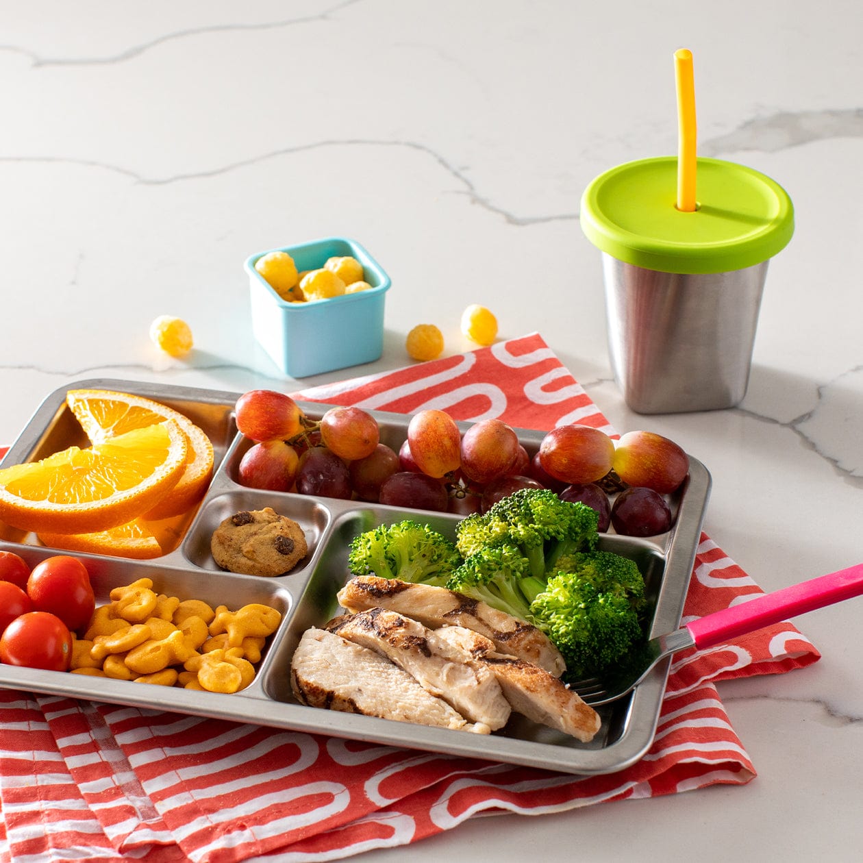 Cup with green silicone lid and yellow straw featured next to Stainless Steel stray with orange slice, grapes, chicken and broccoli, cheddar fish snacks, tomatos and mini cookie. 