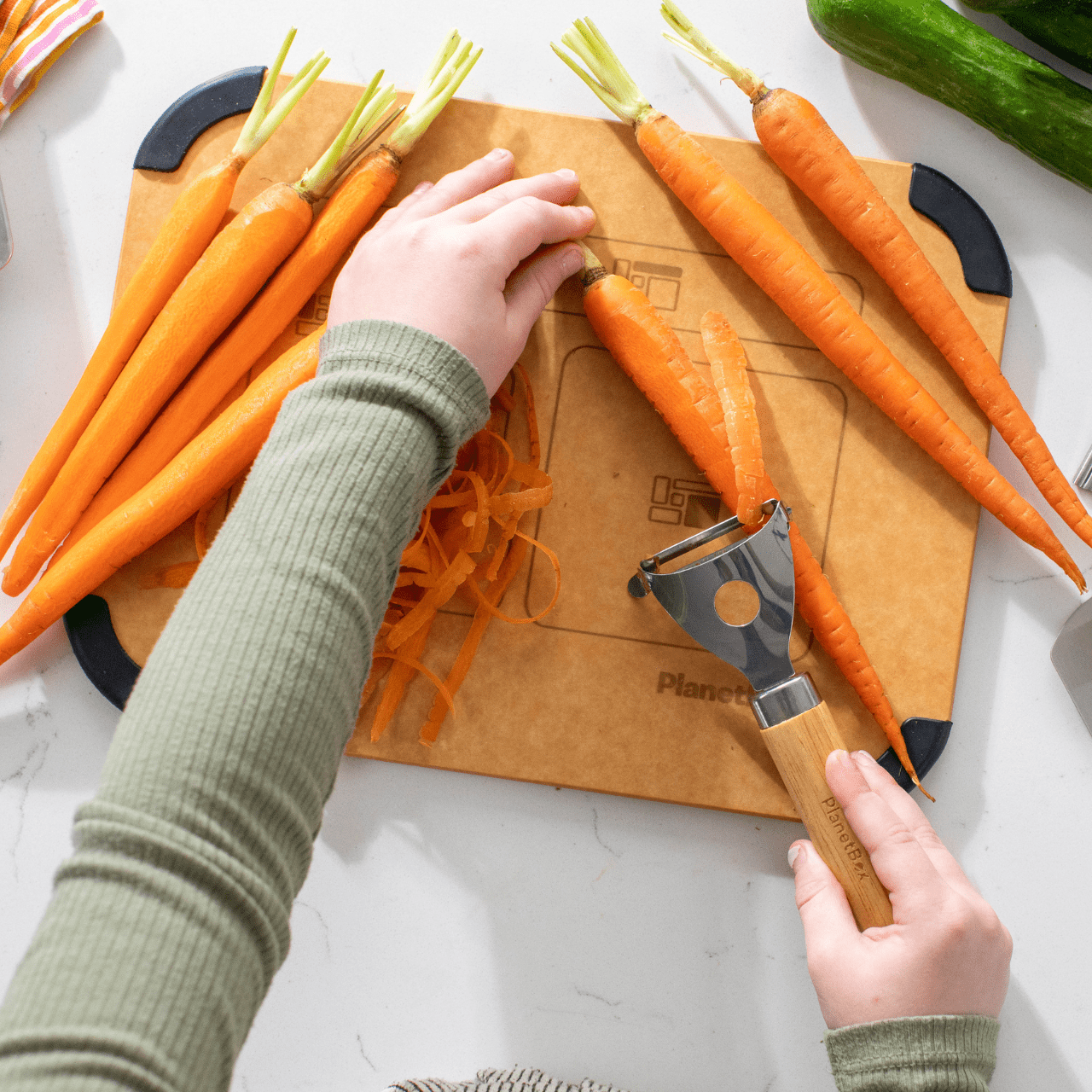 Child using vegetable peeler from Prep to Pack Three Piece Set on fresh carrots atop Prep to Pack cutting board on white counter top.