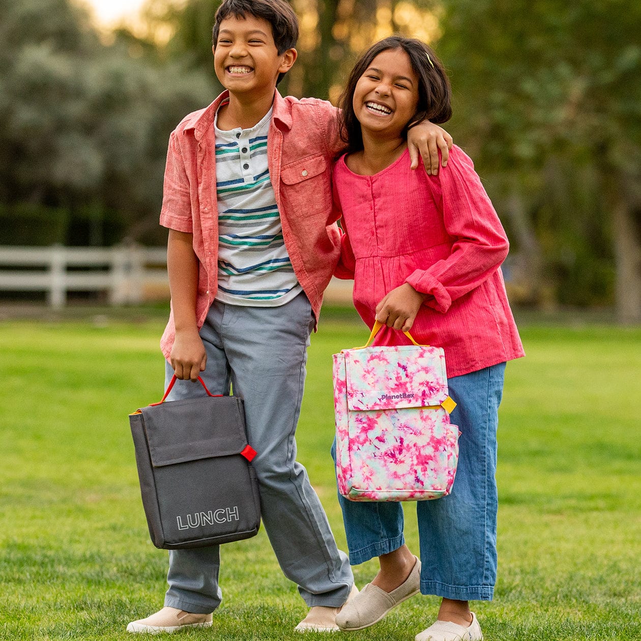 Two children laughing and holding lunchboxes in a field.