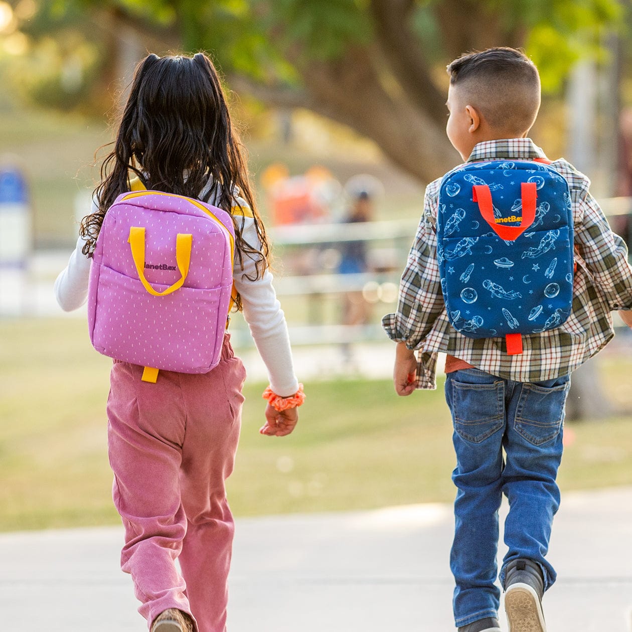 Two children walking in park wearing lunch totes on their backs.